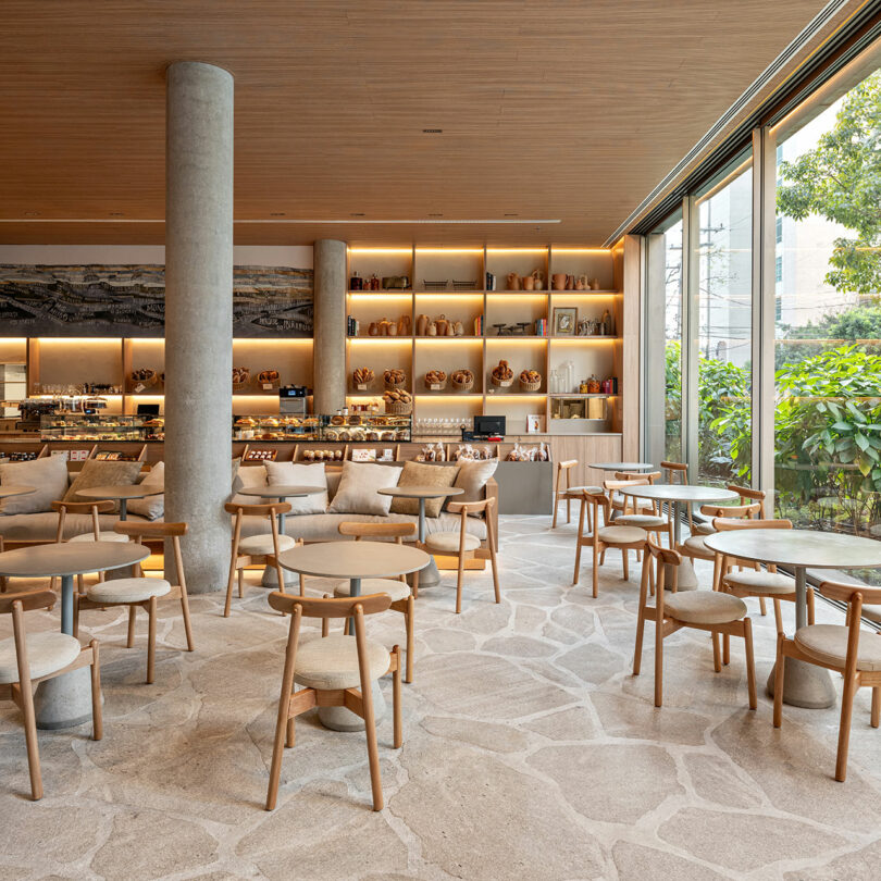Modern cafe interior with wooden tables and chairs, open shelves displaying pottery, large windows, and natural light illuminating the neutral-toned space.