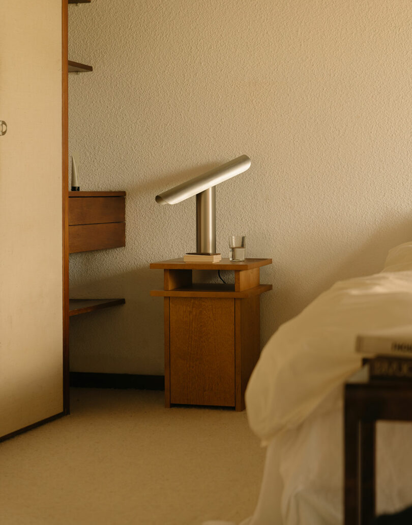 A bedside table with a modern silver lamp and a glass of water next to a bed with white bedding in a softly lit room