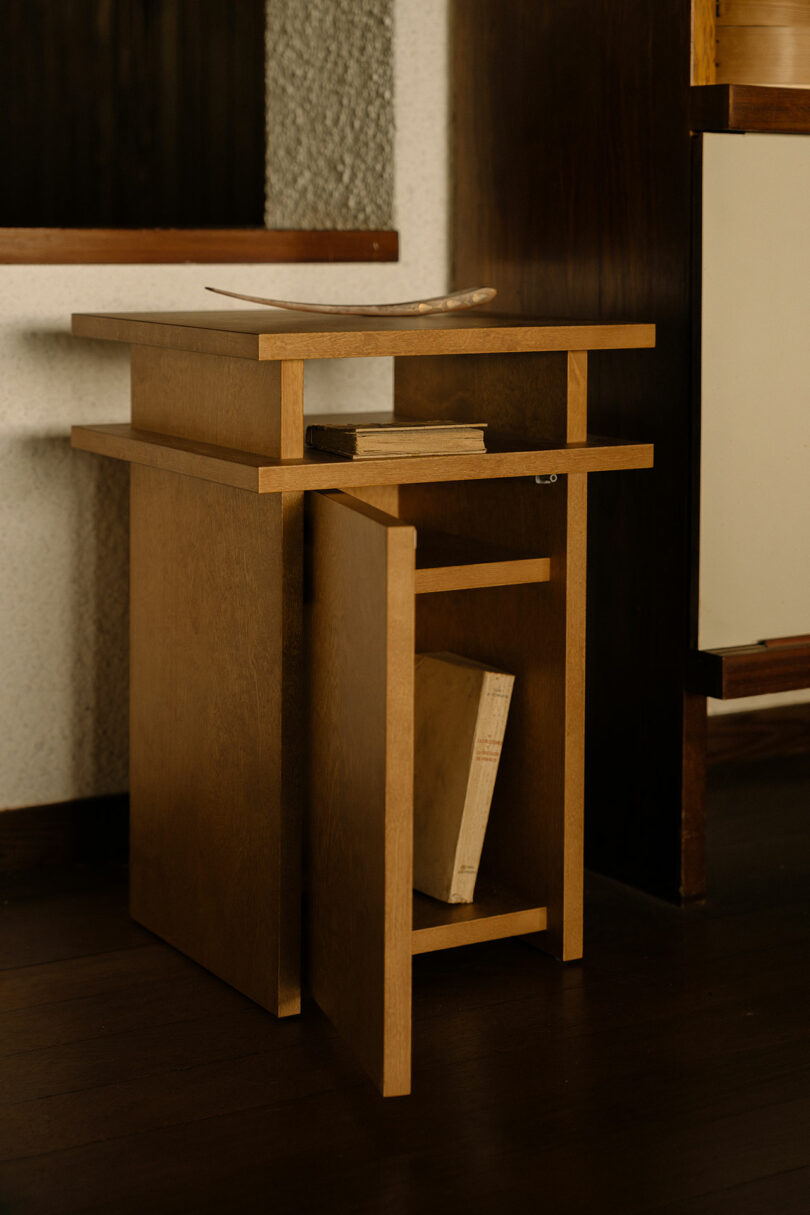A small wooden side table with an open lower compartment holding two books, placed on a dark wood floor next to a wall.