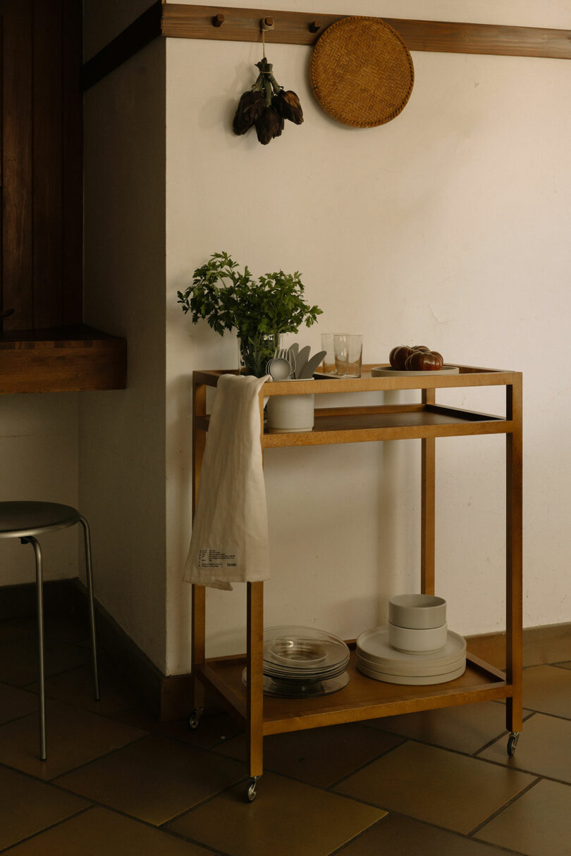 A wooden serving cart with plates, glasses, a plant, and a white towel is positioned against a white wall in a kitchen area. A wicker tray and dried herbs hang above