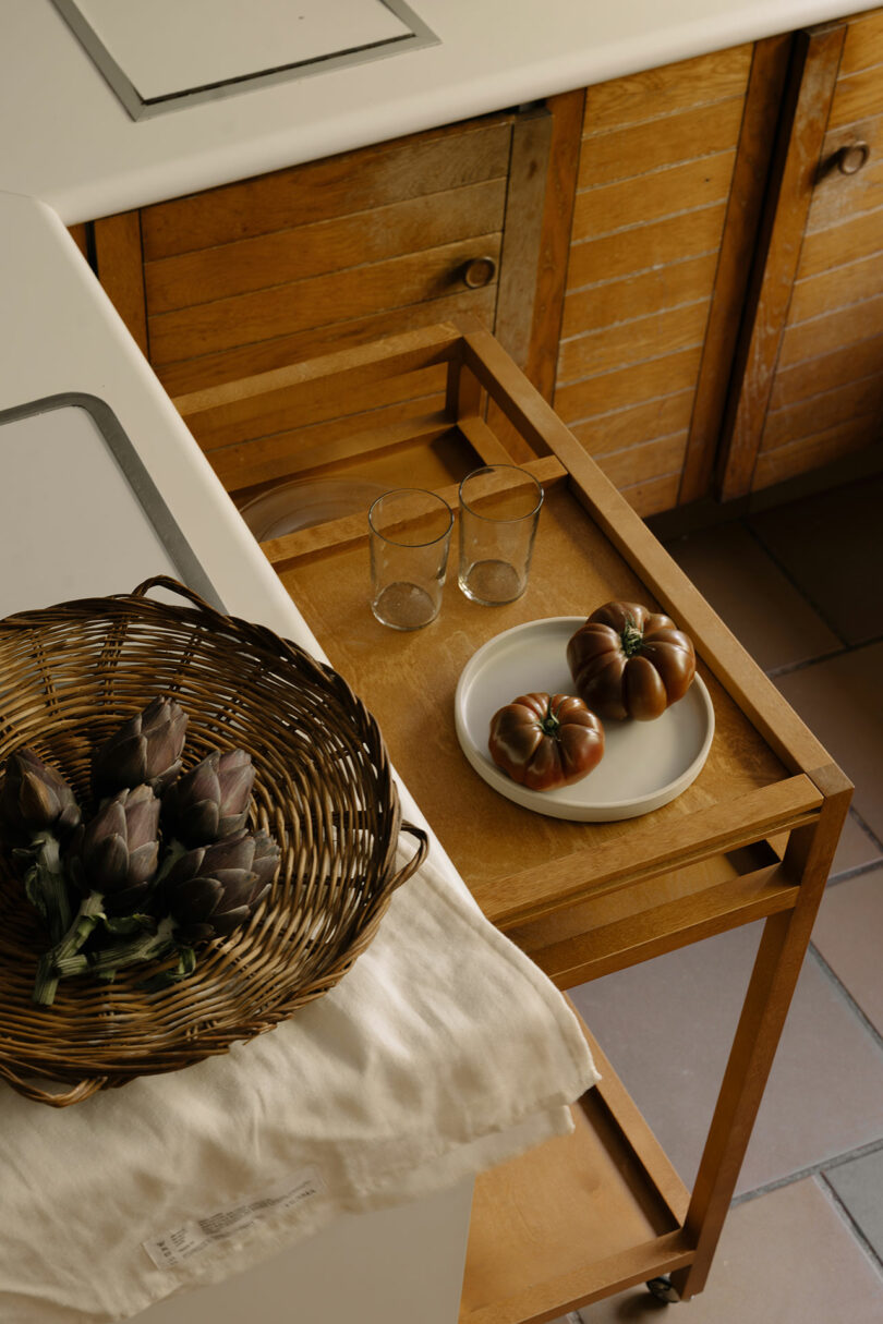 A wooden kitchen cart holds a white plate with tomatoes and two glasses. Nearby, a wicker basket with artichokes rests on a white cloth by the counter