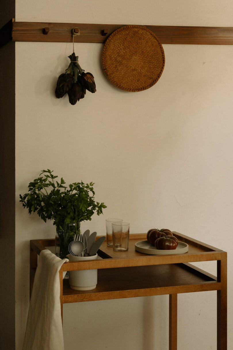 A wooden cart holds a plant, kitchen utensils, and a plate with tomatoes; above it, a woven basket and dried eggplants hang on the wall