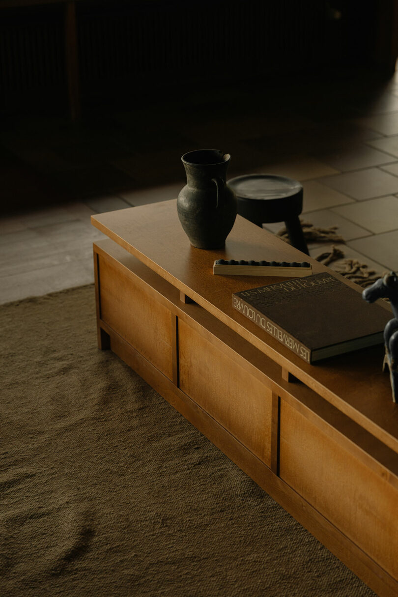 A wooden coffee table with a ceramic pitcher, a closed book, and a striped object on top, placed on a beige rug in a softly lit room