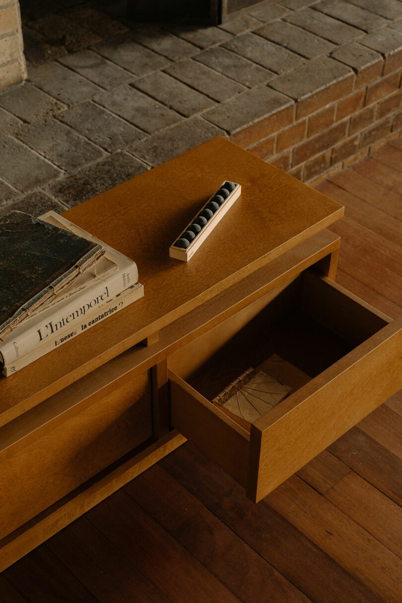 A wooden table with an open drawer sits on a wood floor; books and a small box with black spheres rest on top, in front of a brick fireplace