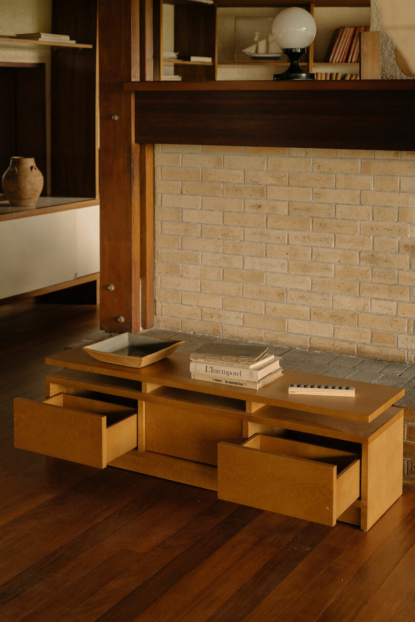 A low wooden table with three open drawers sits in front of a brick fireplace, holding books and trays, in a room with wood flooring and mid-century decor