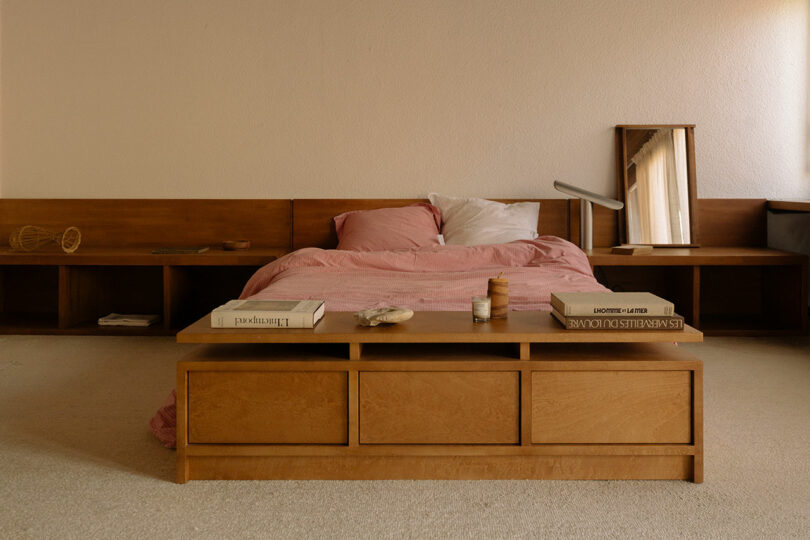 A minimalist bedroom with a wooden bed frame, pink bedding, a bedside lamp, a mirror, and books on a wooden bench at the foot of the bed