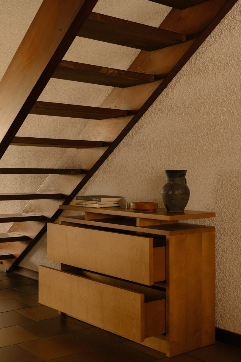 A wooden sideboard with open drawers sits beneath a floating wooden staircase. On top of the sideboard are books, a bowl, and a decorative vase