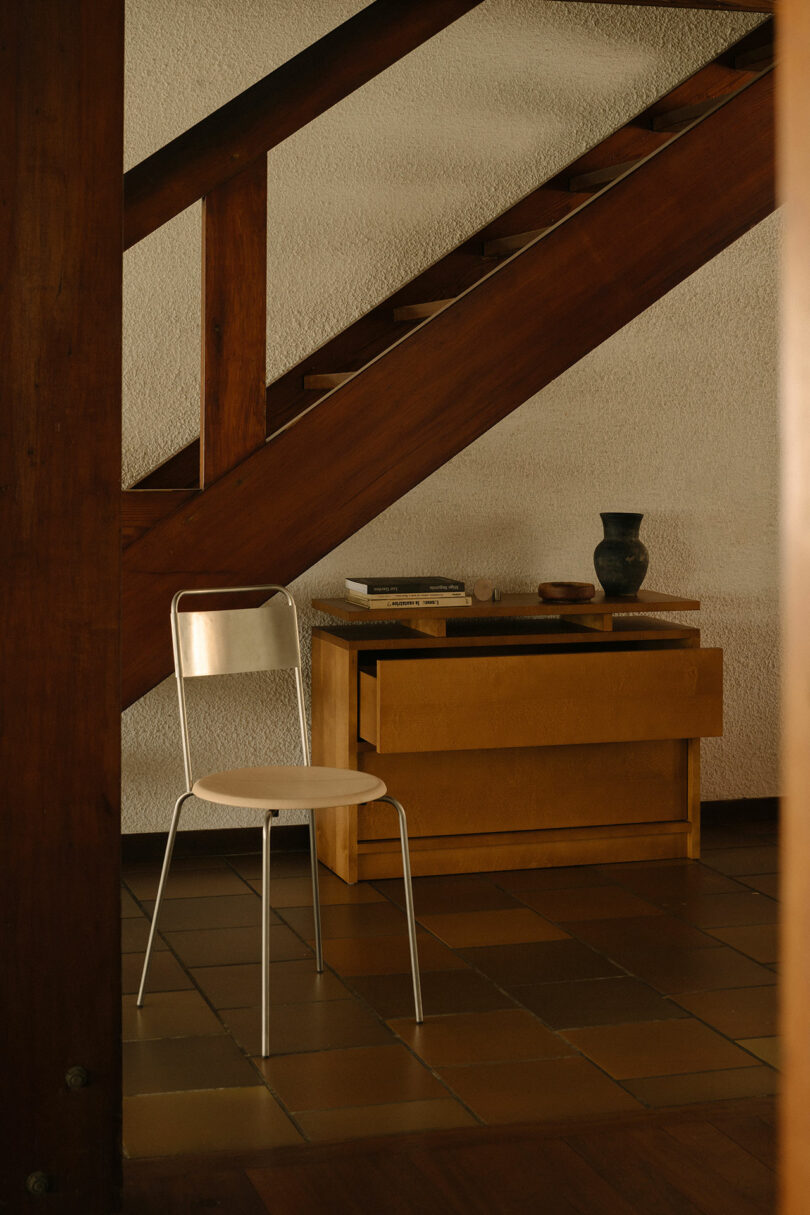 A metal chair with a light wooden seat is placed under a wooden staircase beside a small wooden cabinet with books and a dark vase on top