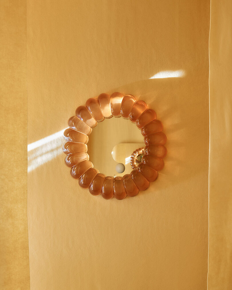 A round mirror with a translucent amber frame is mounted on a yellow wall, reflecting a white sphere and soft light streaming in from the left