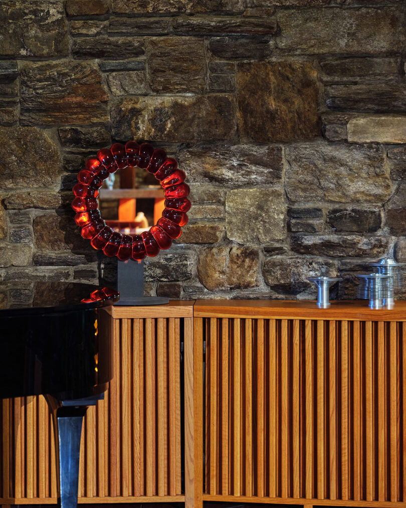 A circular red glass-framed mirror sits on a wooden cabinet against a stone wall, with metal objects and the corner of a black piano nearby