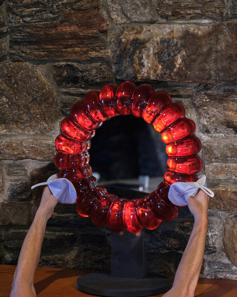 Person holding a round, red-glass sculptural mirror with both hands in front of a stone wall