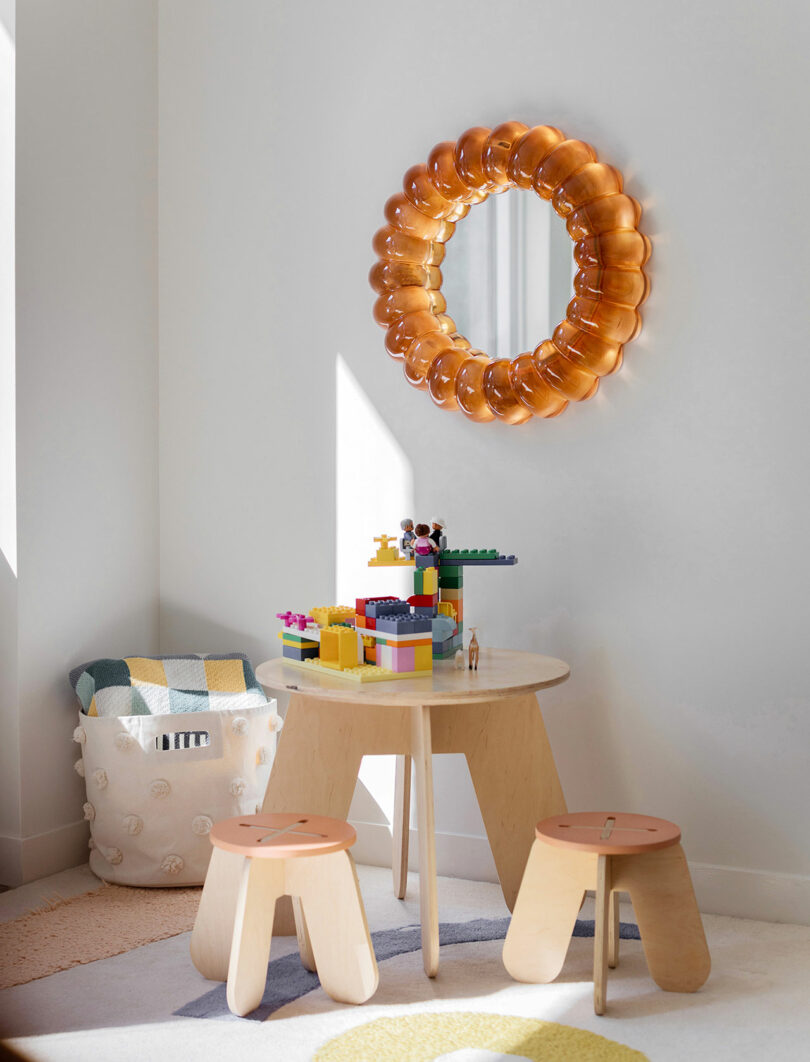 A small play area with a round wooden table, two stools, building blocks on the table, a knitted basket, and a circular amber mirror on the wall