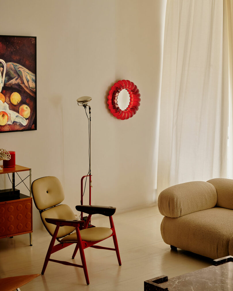 A modern living room with a beige armchair, red-accented chair, red wall mirror, floor lamp, abstract painting, and light curtains covering a tall window