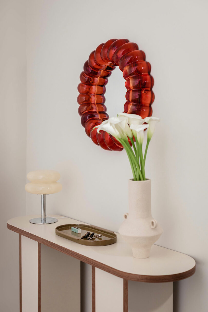 A modern console table with a vase of white flowers, a small tray with items, a sculptural lamp, and a circular red wall mirror above