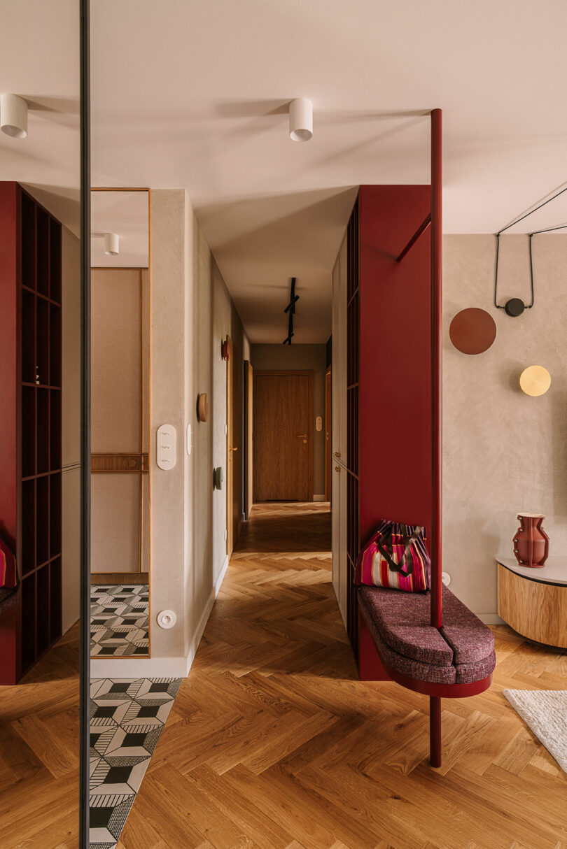 Modern hallway with wooden herringbone floor, maroon seating area, geometric tile, wall-mounted art, and a wooden door at the end. A mirror reflects part of the space.