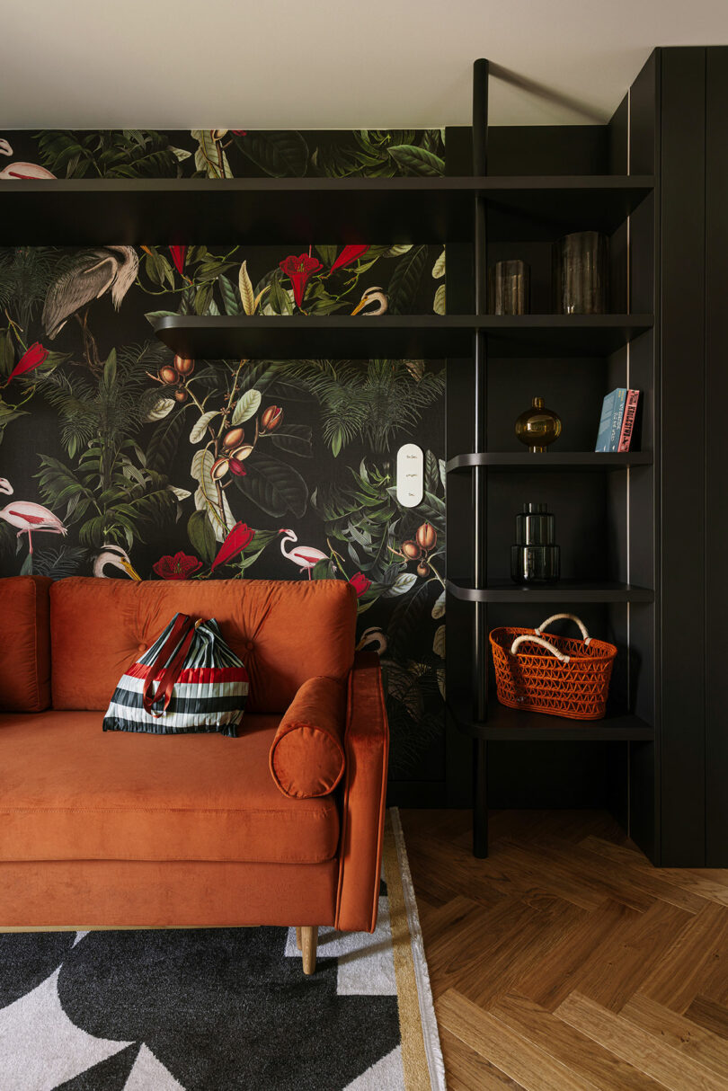 A rust-colored sofa sits in front of a wall with a dark botanical and bird print wallpaper, next to black open shelving with decor items and books.