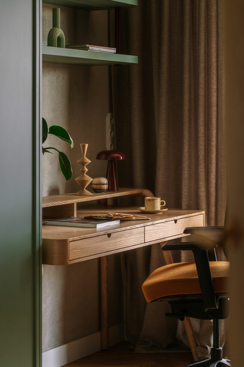 A wooden desk with a notepad, pen, and mug sits near a window with brown curtains; a chair and decorative objects are arranged on and above the desk.