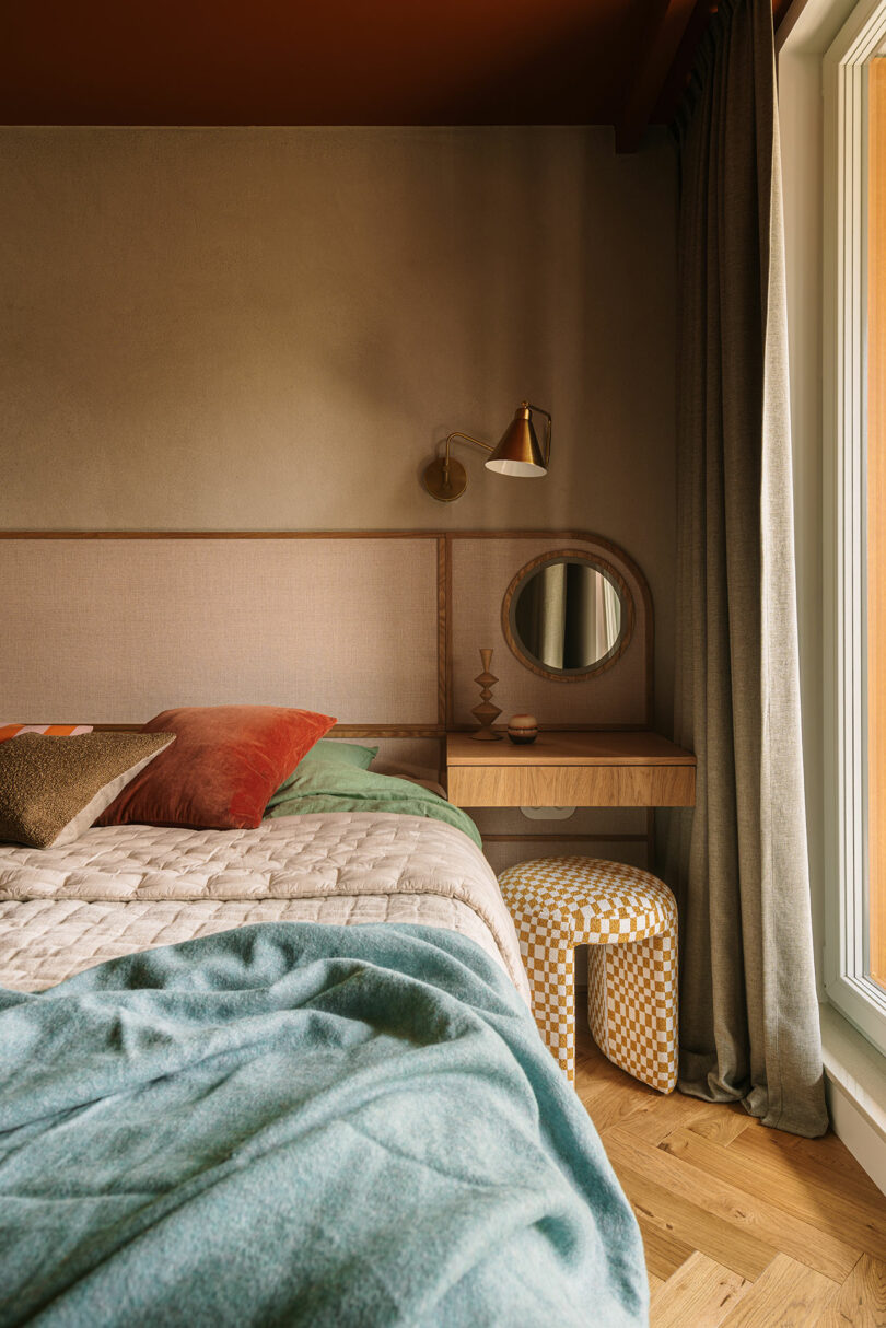 A cozy bedroom corner features a bed with colorful pillows and a blanket, a small wooden vanity with a round mirror, a checkered stool, and a window with curtains.