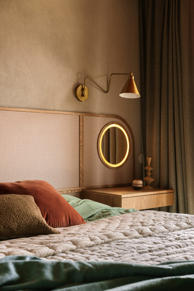 A neatly made bed with pillows sits next to a wooden nightstand with a round, illuminated mirror, geometric decor, and a gold wall lamp above. Dark curtains are in the background.