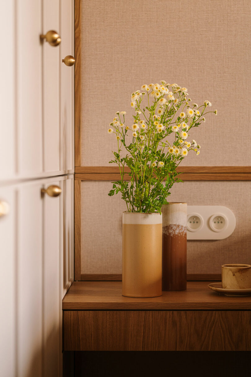 A ceramic vase with white and yellow daisies sits on a wooden table next to a smaller vase and a cup; built-in cabinets and wall outlets are visible in the background.