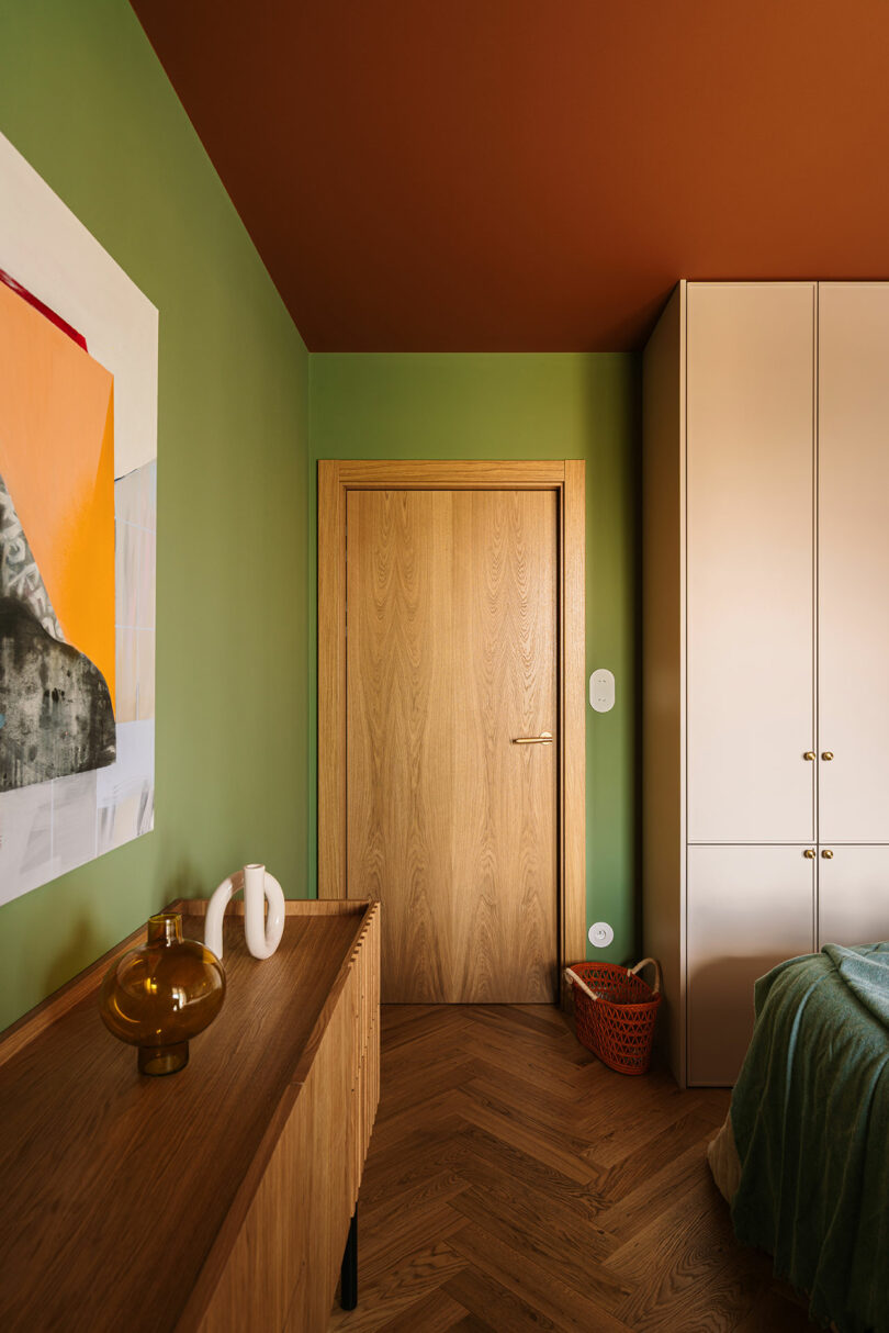 A room with green walls, a wooden door, a wooden sideboard with decorative objects, a white wardrobe, a bed with a green blanket, and an orange ceiling.