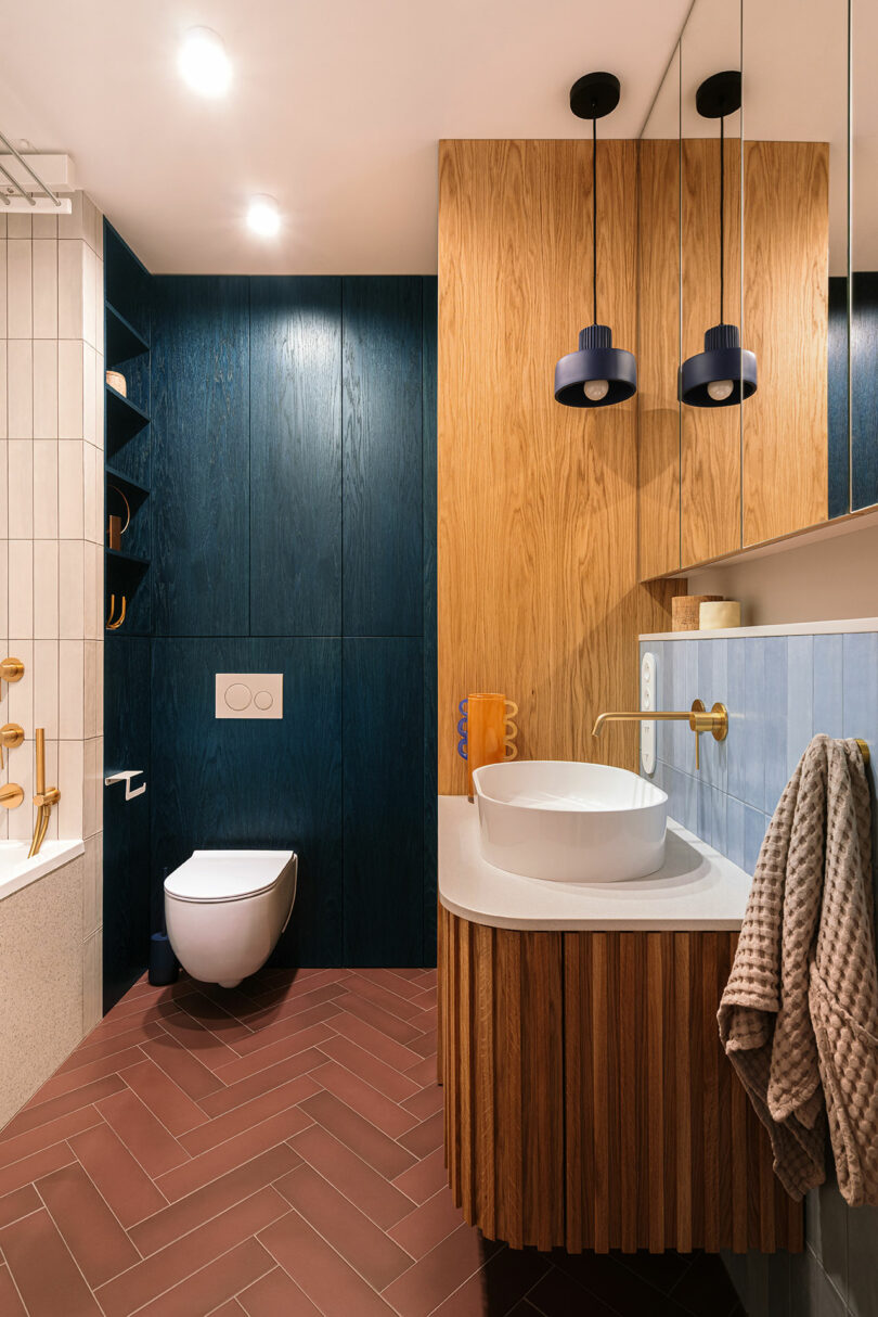 Modern bathroom with a wall-mounted toilet, vessel sink, wood and dark blue paneling, pendant lights, and gold fixtures. Towel hangs on a hook beside the sink.