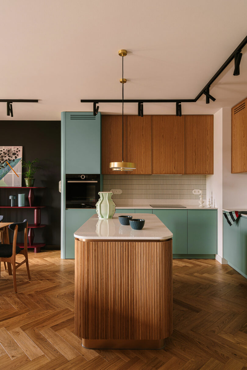 Modern kitchen with light wood and green cabinets, a marble-topped island, pendant lighting, and herringbone wood flooring.
