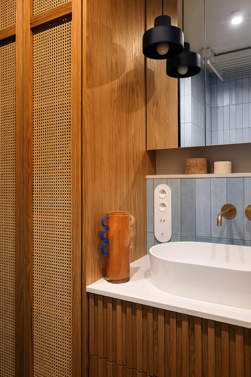 Modern bathroom with wood paneling, a round white basin sink, blue tile backsplash, brass fixtures, a brown vase with blue handle, and a black pendant light.