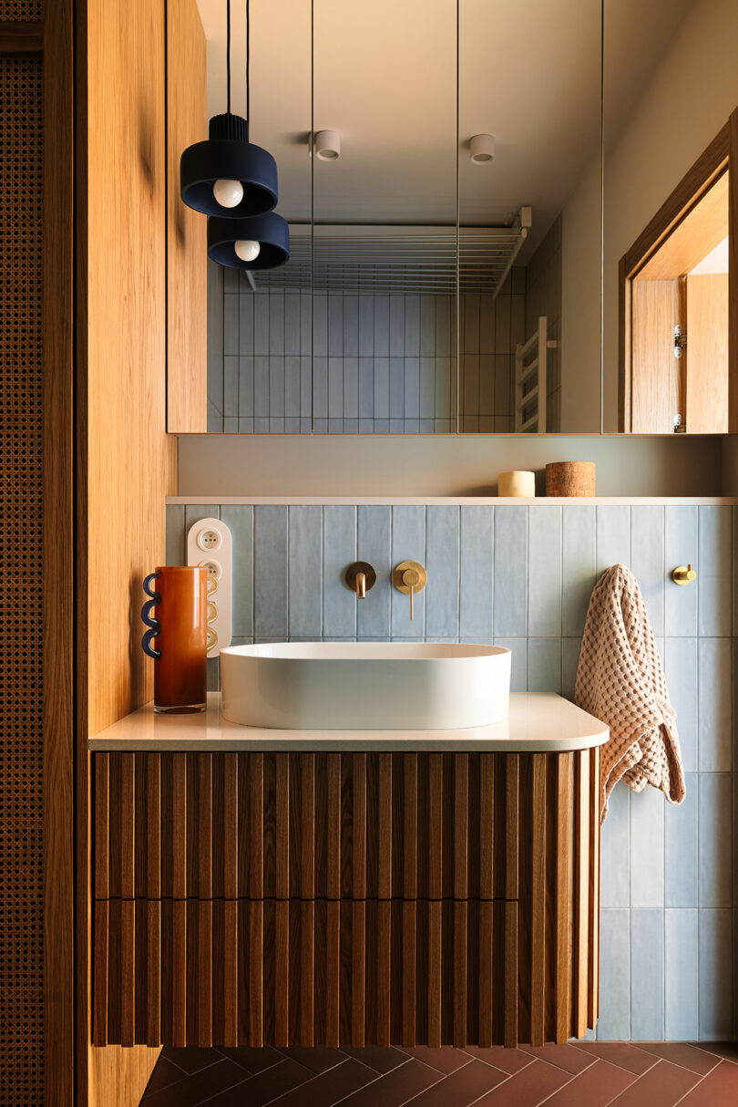 Modern bathroom with a wood vanity, vessel sink, wall-mounted faucet, light blue tiled backsplash, large mirrored cabinet, and pendant lights. A towel hangs on the wall.