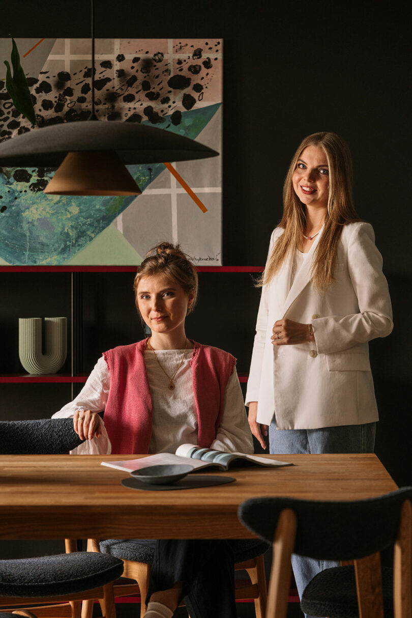 Two women pose in a modern room; one is seated at a table with an open magazine, and the other stands beside her. Contemporary art and shelves are visible in the background.