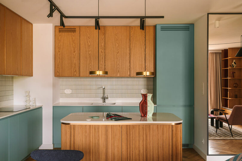 Modern kitchen with wood cabinets, light blue lower cabinets, white countertop, two pendant lights, and decorative vases on the island. A living room is visible in the background.