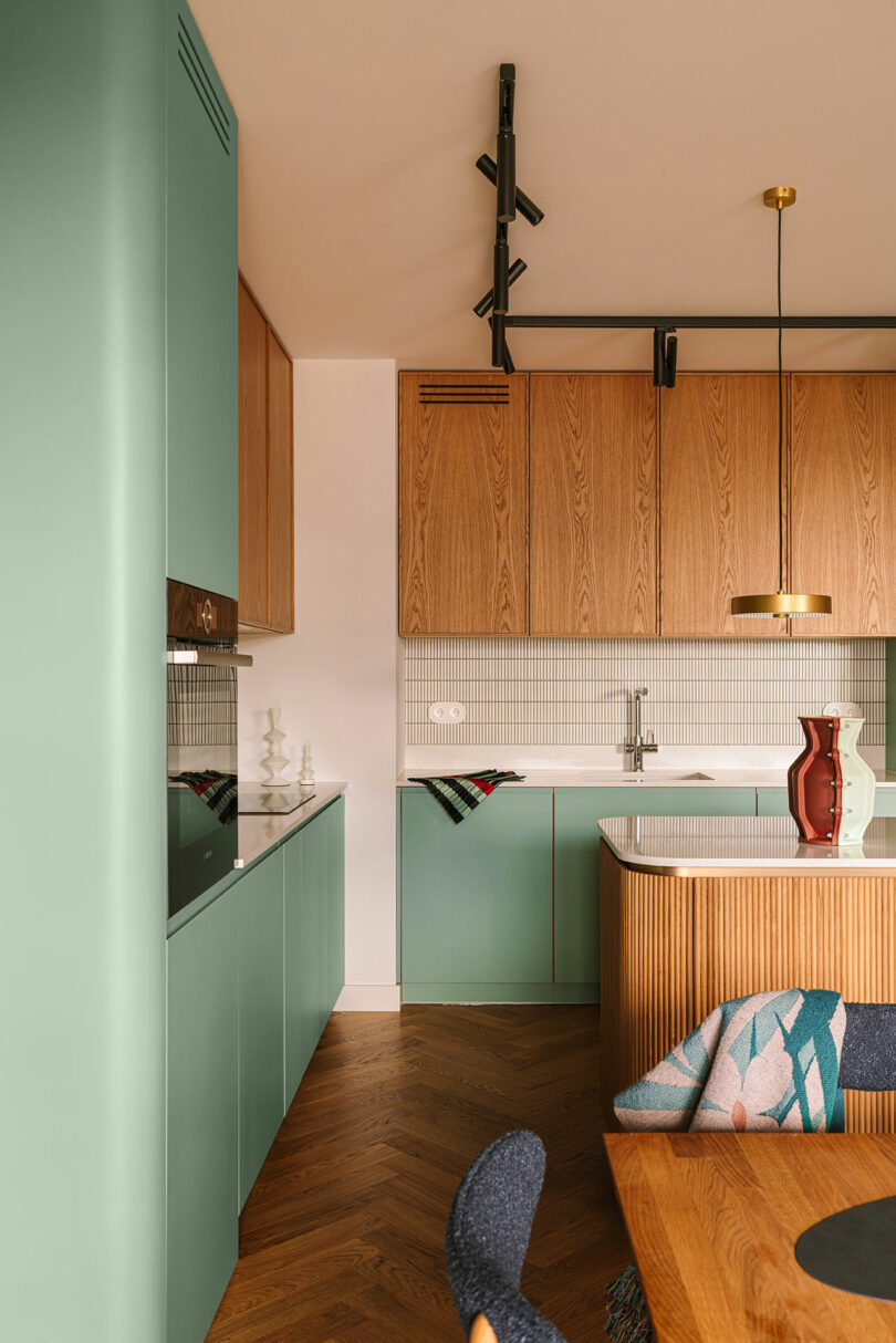 Modern kitchen with mint green cabinets, wooden upper cabinets, a white backsplash, gold pendant light, and decorative items on the counter and table.
