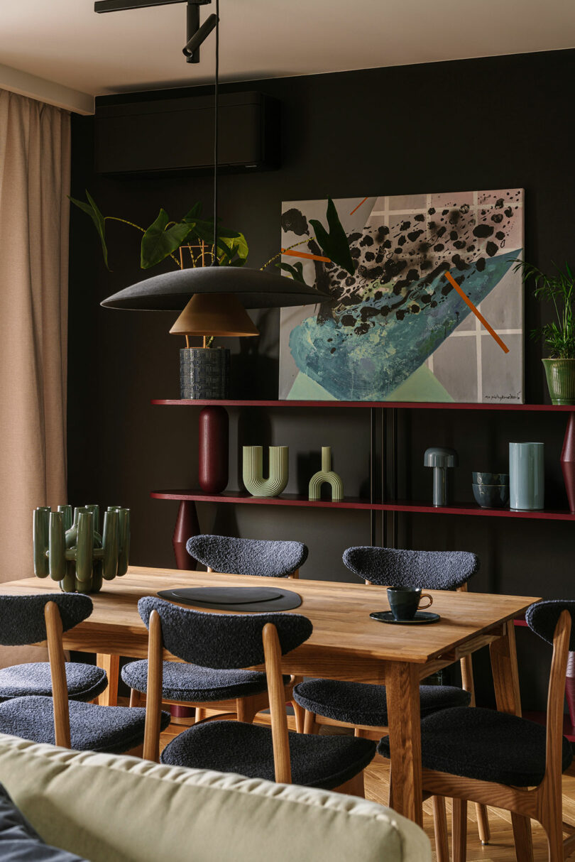 A modern dining room with a wooden table, six upholstered chairs, green glassware, a black pendant light, art on a dark wall, and decorative vases on red shelves.