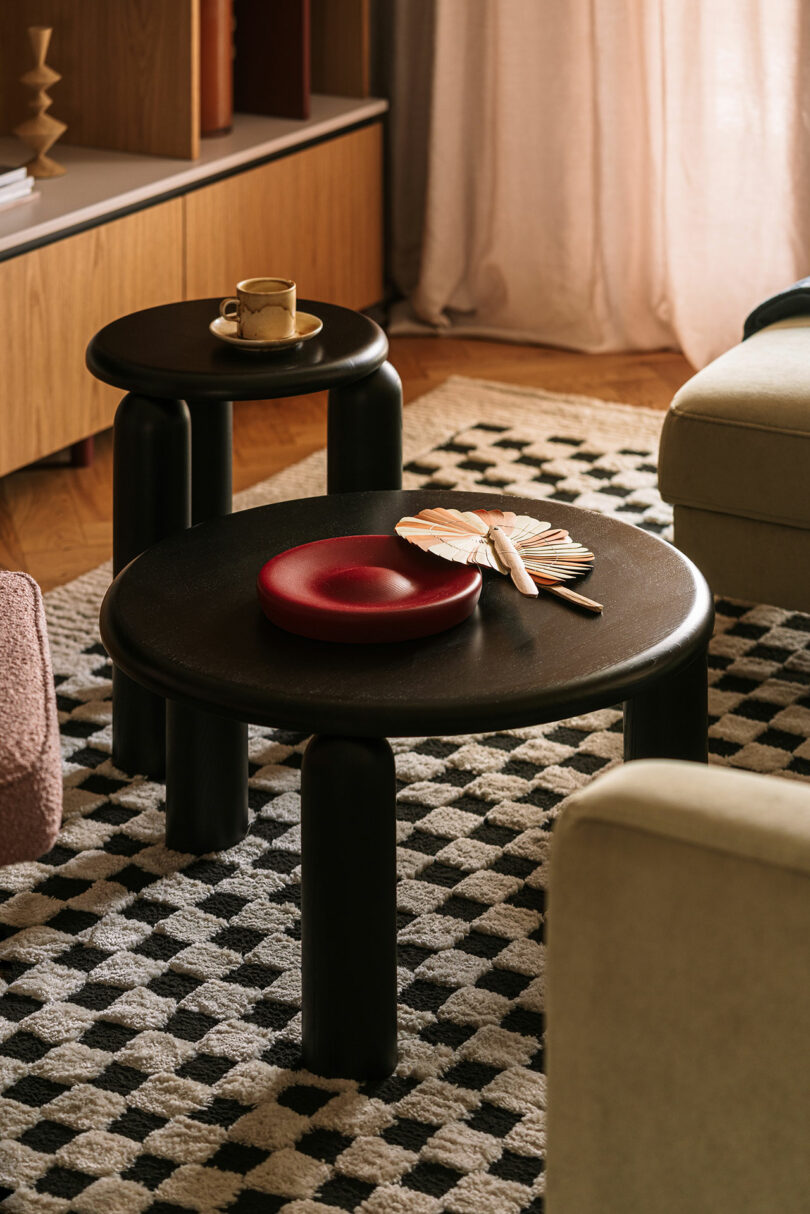 Two round black tables on a black-and-white checkered rug, with a cup on one table and a red plate with fans on the other, surrounded by modern furniture and soft lighting.