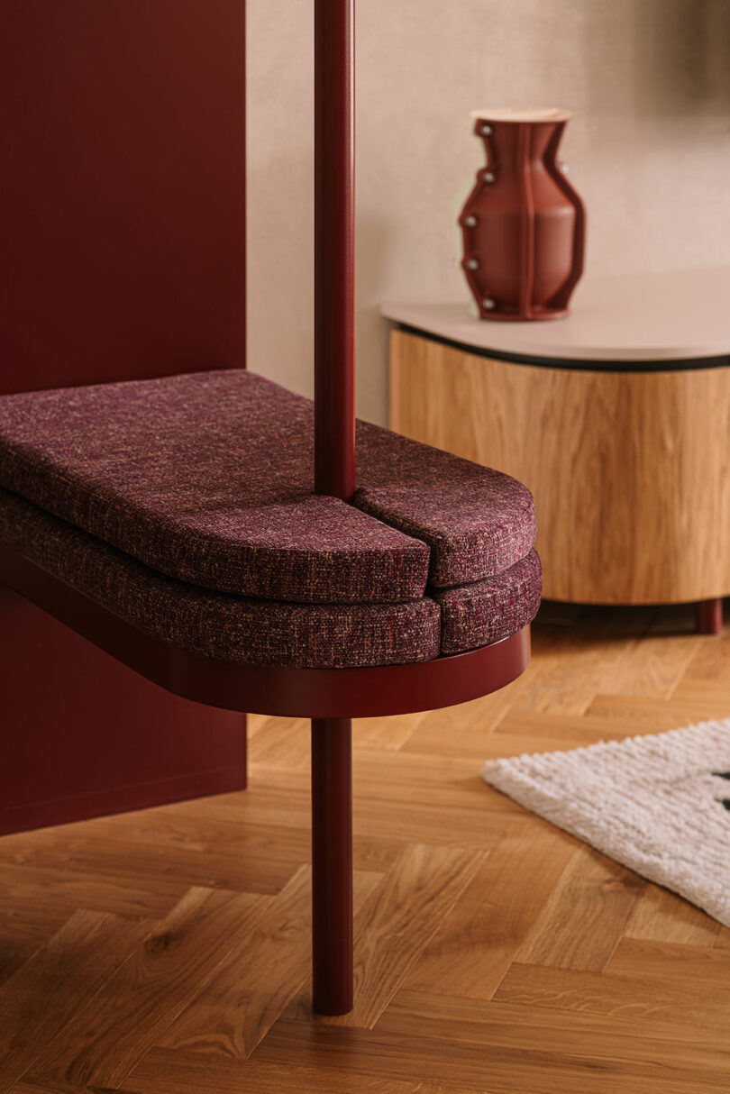 A modern interior with a burgundy cushioned bench attached to a vertical pole, a wooden cabinet with a vase, and a textured rug on a herringbone wood floor.