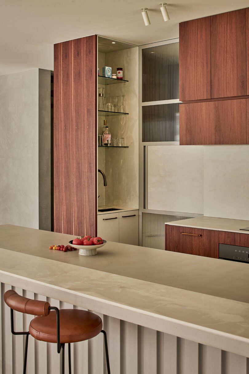 Modern kitchen with wood cabinets, open shelves with glassware, a built-in sink, and a light countertop island with a brown leather bar stool and a bowl of fruit.