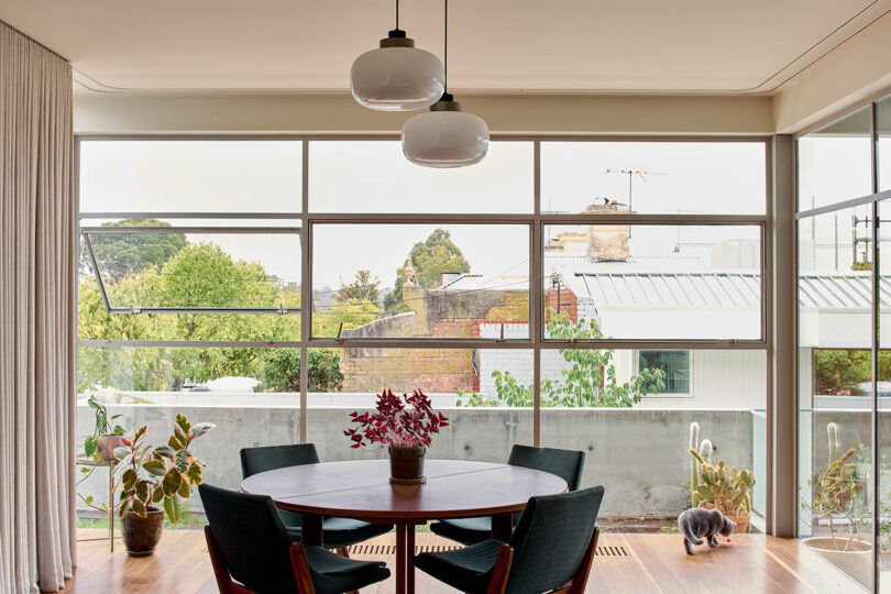 A dining area with a round table, five chairs, potted plants, large windows, and a cat walking on a wooden floor near the balcony.