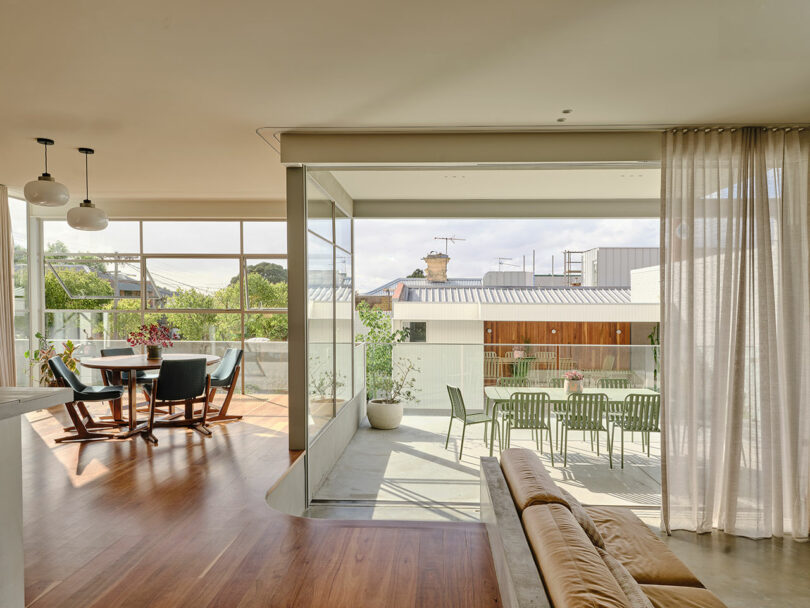 Modern open-plan living and dining area with wooden floors, large windows, and sliding glass doors leading to an outdoor patio with green chairs and a table.