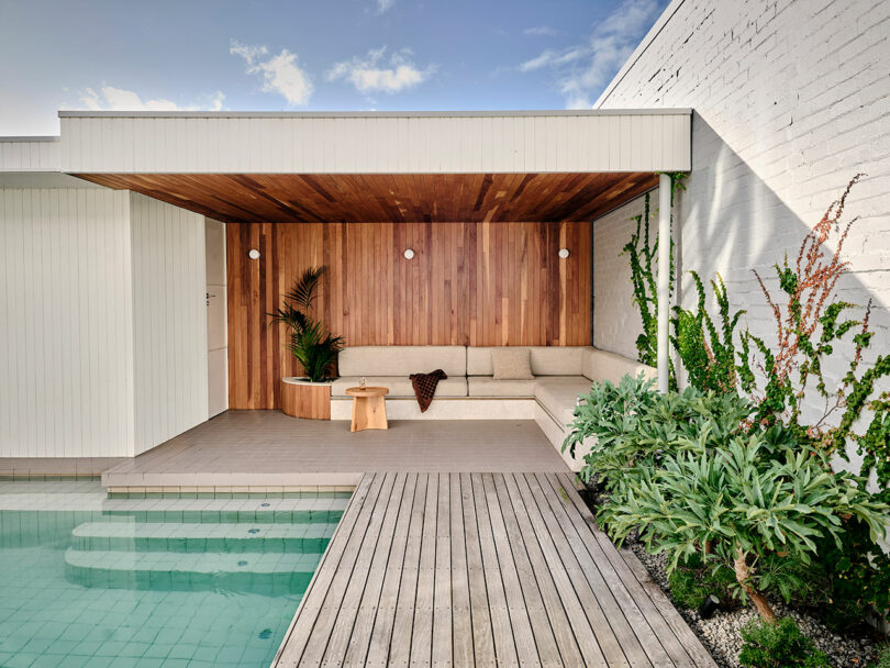 Modern poolside lounge area with a wooden deck, beige sectional sofa, small wooden stool, potted plants, and a partially covered ceiling next to a white brick wall.