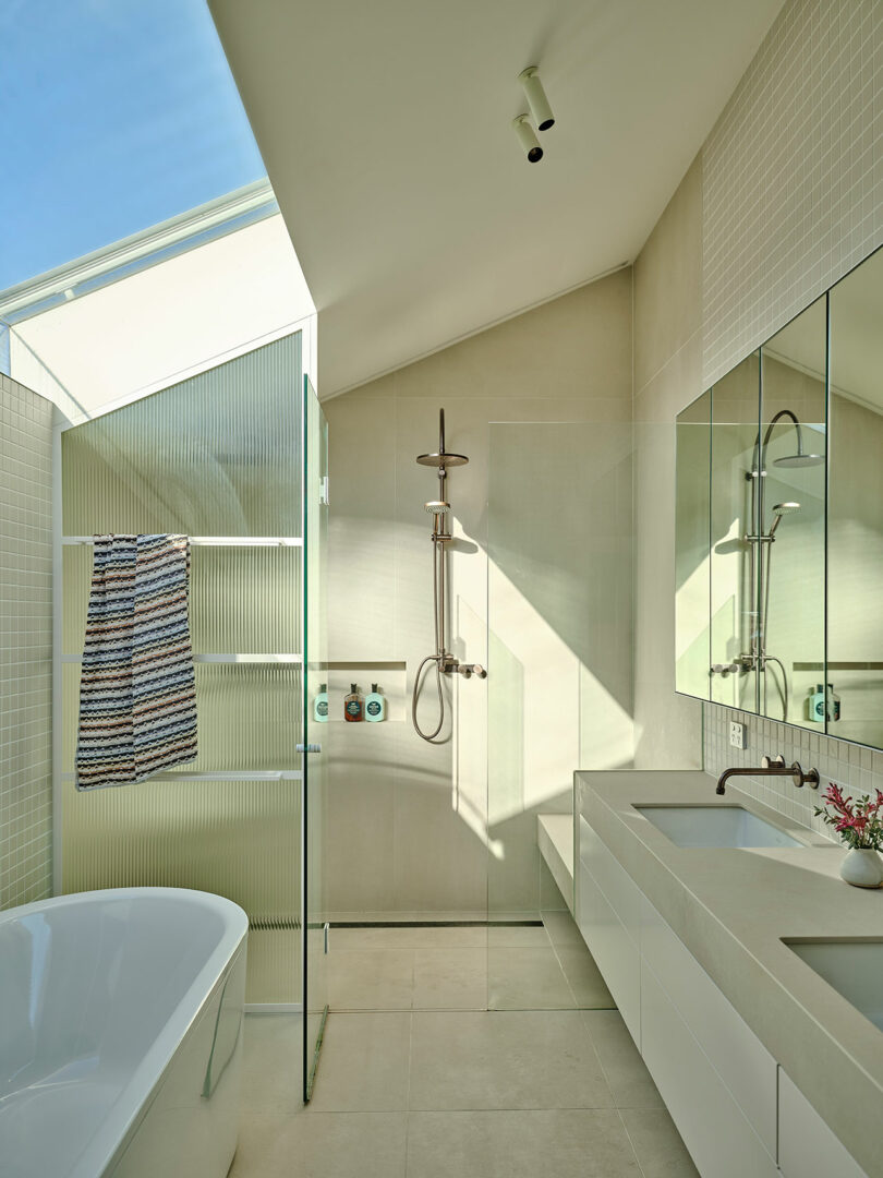 Modern bathroom with a freestanding tub, glass-enclosed shower, wall-mounted fixtures, large mirrors, and neutral-toned tile under a slanted ceiling with natural light.