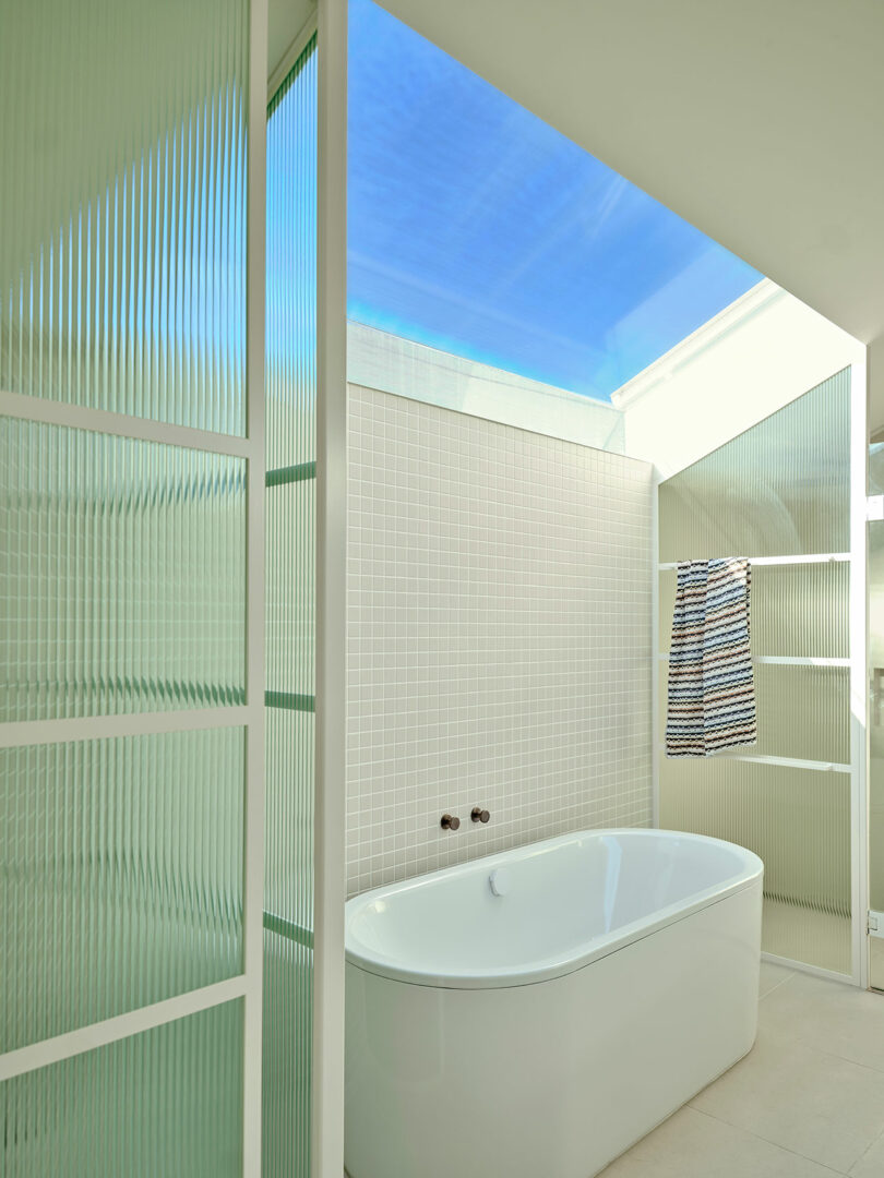 A modern bathroom with a white freestanding tub, frosted glass panels, a skylight above, tiled walls, and a towel hanging on open shelving.