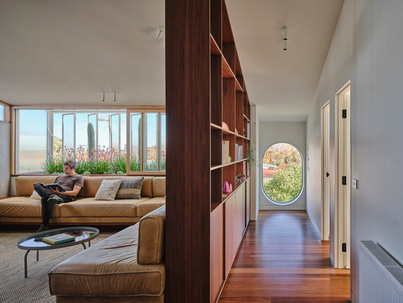 Spacious modern living room with tan sectional sofa, built-in wooden shelving, large windows, and a hallway featuring a round window. A person is sitting on the sofa reading.