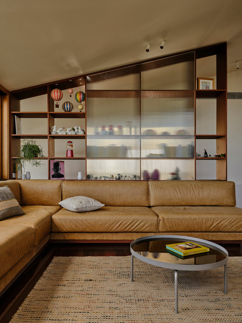 A modern living room with a tan leather sectional sofa, round glass coffee table, and open shelves displaying decor items, including model hot air balloons and small sculptures.