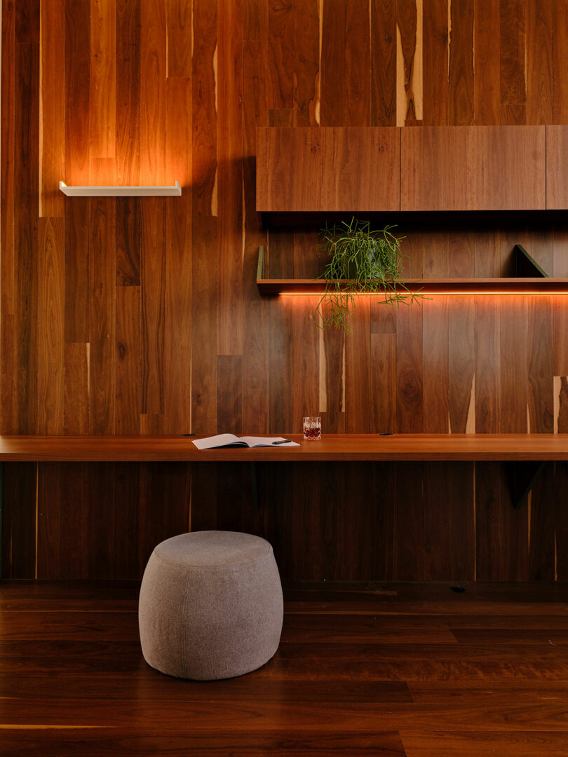 A modern wooden wall with built-in shelves, a bench with an open book and drink, a potted plant, and a gray round ottoman in front. Warm lighting creates a cozy ambiance.