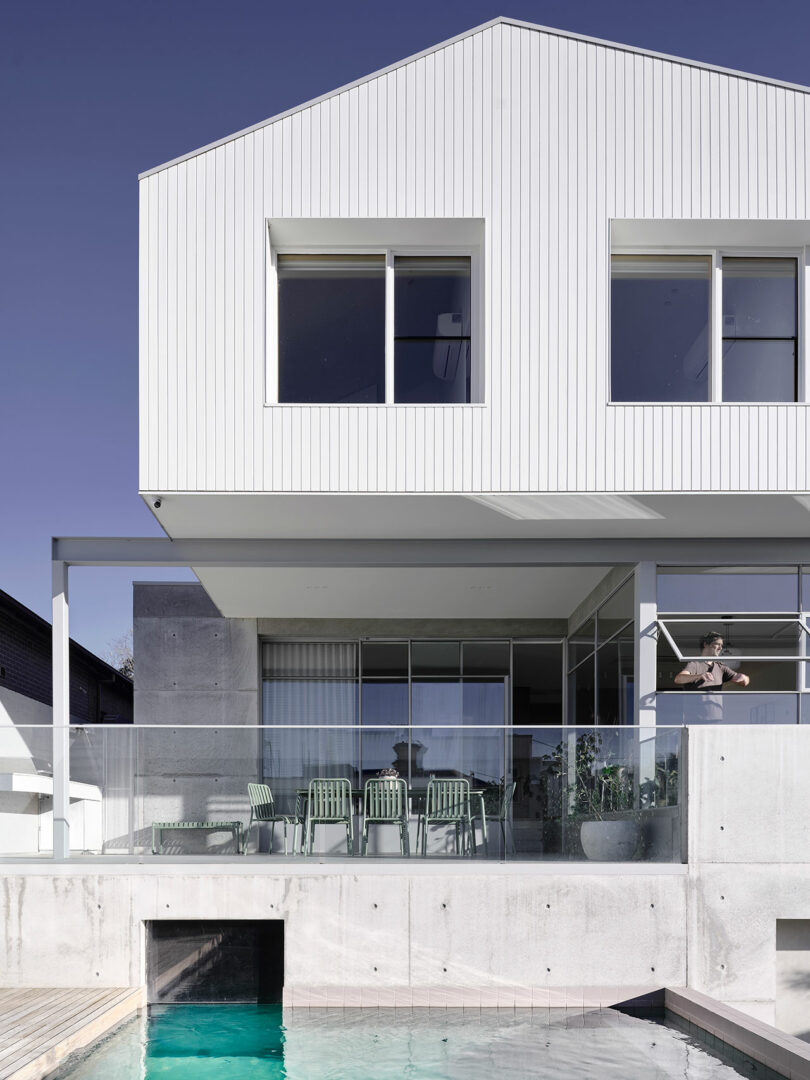 Modern two-story white house with large windows, a covered patio with green chairs, glass railing, and a swimming pool in the foreground under a clear sky.