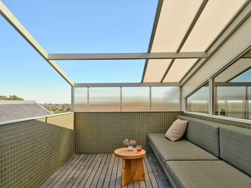 A modern outdoor patio with a green-tiled wall, gray cushioned bench, wooden deck, small round wooden table, and a beige pillow under a partially covered roof.