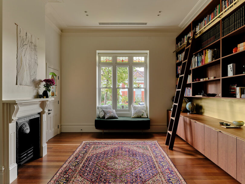 A living room with a large patterned rug, built-in bookshelf with ladder, window seat, fireplace, and wooden floors.
