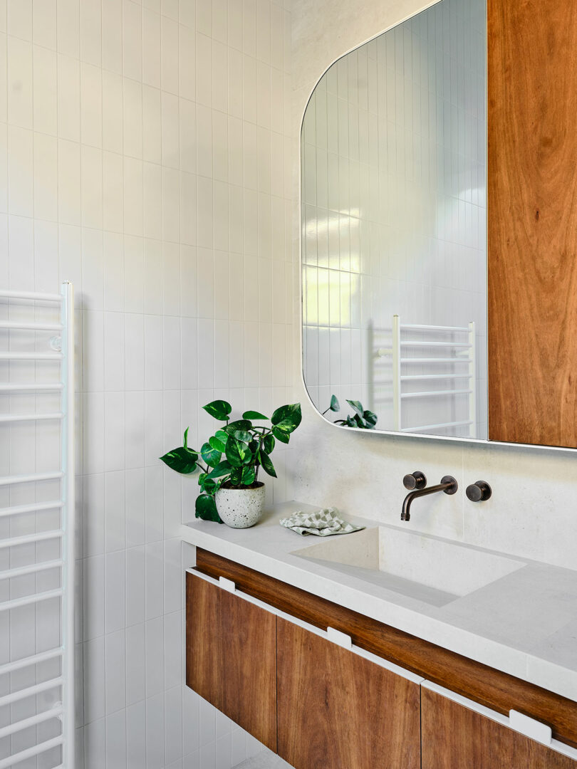Modern bathroom with white tile walls, a large mirror, wooden vanity, built-in sink, wall-mounted faucet, potted plant, and a white towel rack reflected in the mirror.