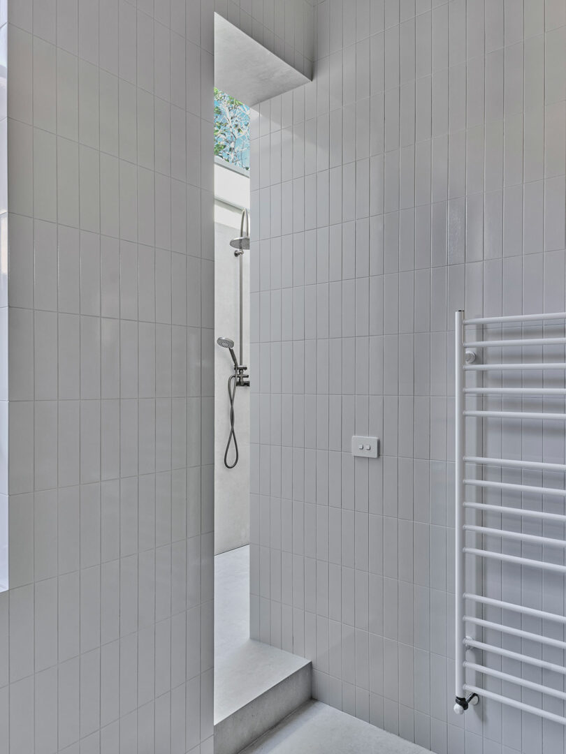 Minimalist white-tiled bathroom with a wall-mounted shower, towel radiator, and a narrow vertical window opening to the outside.