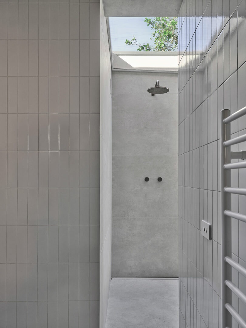 Minimalist shower with light gray tiles, overhead rain shower, and an open skylight showing a tree branch above. A white heated towel rail is on the right wall.